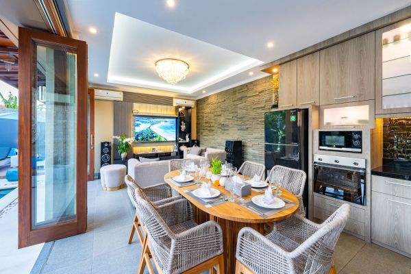 Modern open-plan villa interior with wooden dining table set for meal, rattan chairs, sleek kitchen, TV lounge, and sliding doors to infinity pool. - Bali Villas